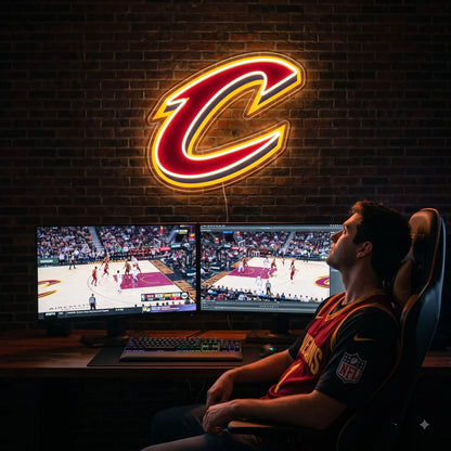 A man sitting at a gaming setup under a large glowing red and gold Cleveland Cavaliers NBA basketball team C logo LED neon sign mounted on a brick wall